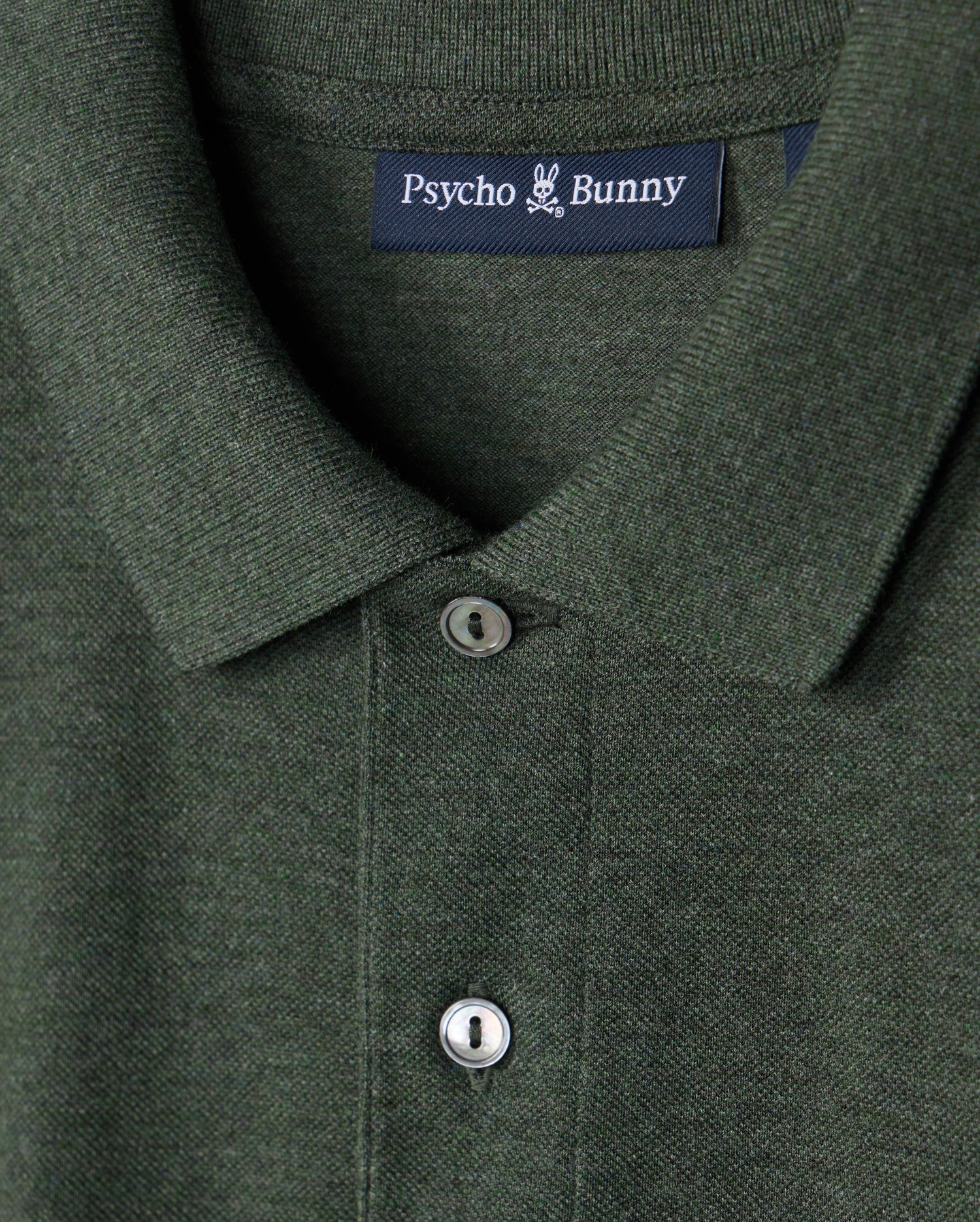 Close-up of a dark green premium pique polo shirt featuring a collar and two buttons. The label displays the brand "Psycho Bunny" in white letters on a dark blue tag, showcasing the high-quality craftsmanship of the Men's James Premium Pique Polo Shirt made from lightweight pima cotton.