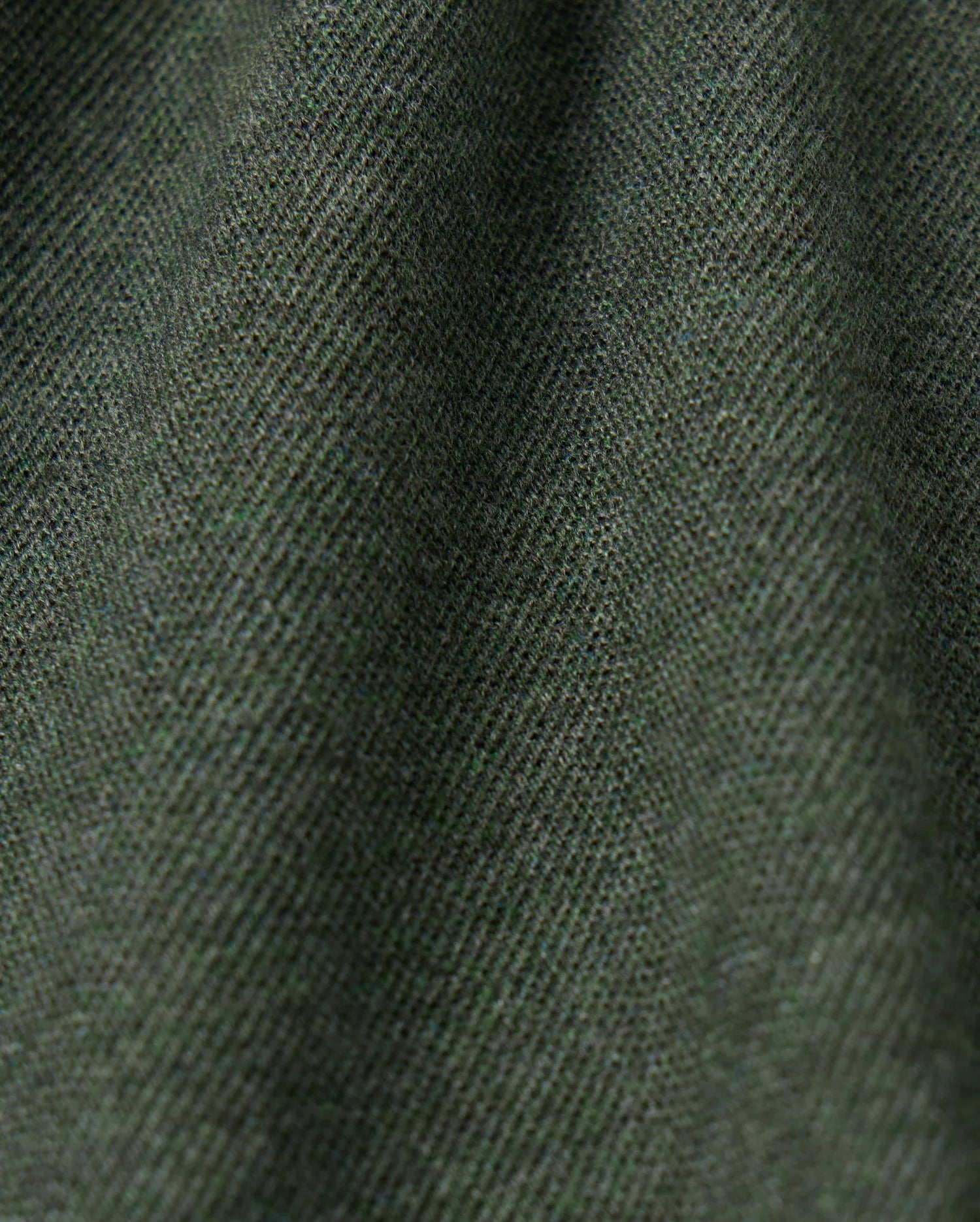 Close-up image of the MENS JAMES PREMIUM PIQUE POLO SHIRT - B6K945D200 by Psycho Bunny, showcasing its dark green premium fabric with a fine diagonal weave pattern that creates a textured appearance. The lighting accentuates the subtle variations in this high-quality polo for men.