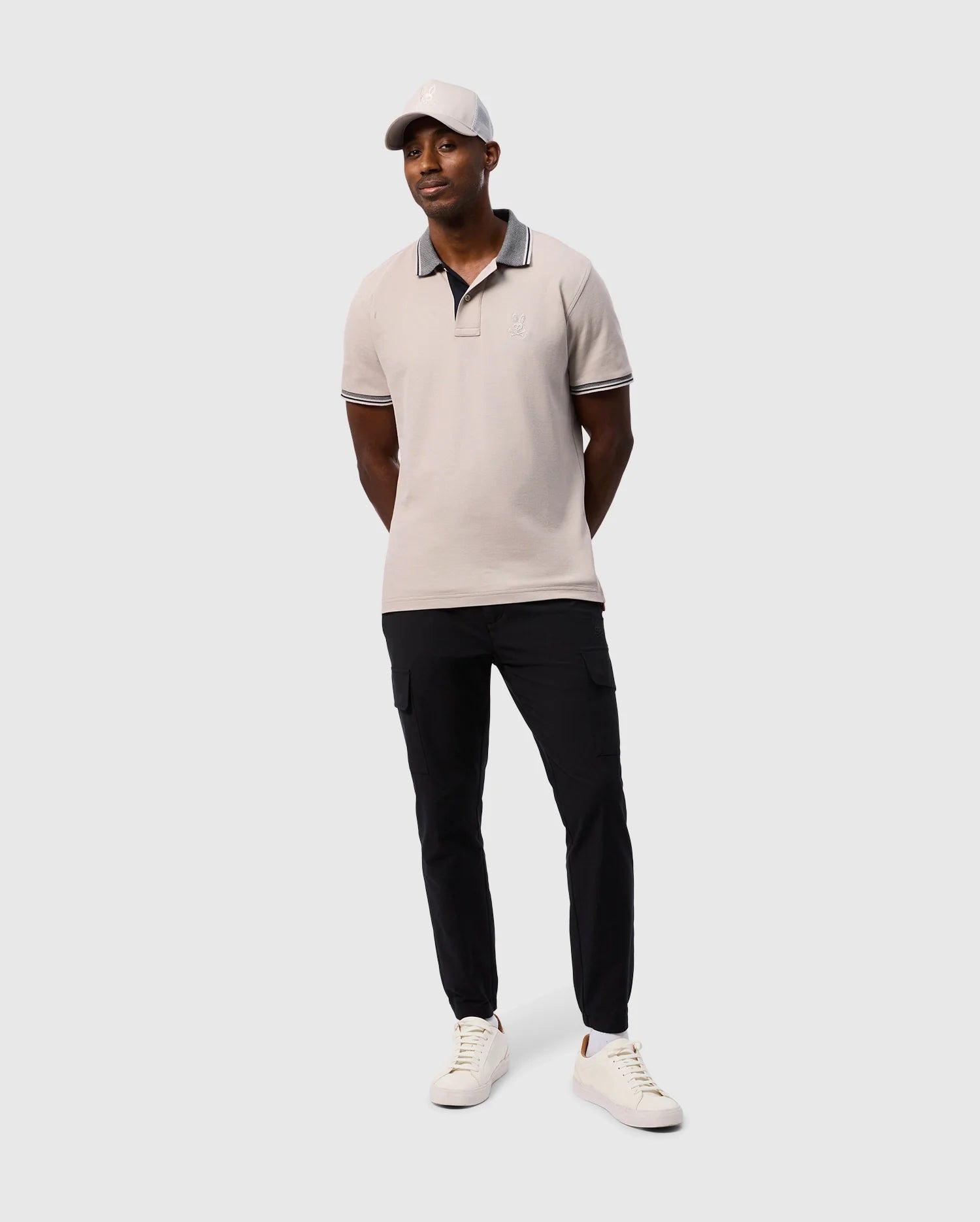 A man stands on a plain background, wearing a Psycho Bunny light grey MENS MALTON PIQUE POLO SHIRT - B6K950D200 with a subtle diamond pique pattern, black cargo pants, white sneakers, and a matching light grey cap. His hands are loosely clasped behind his back, and he is smiling.