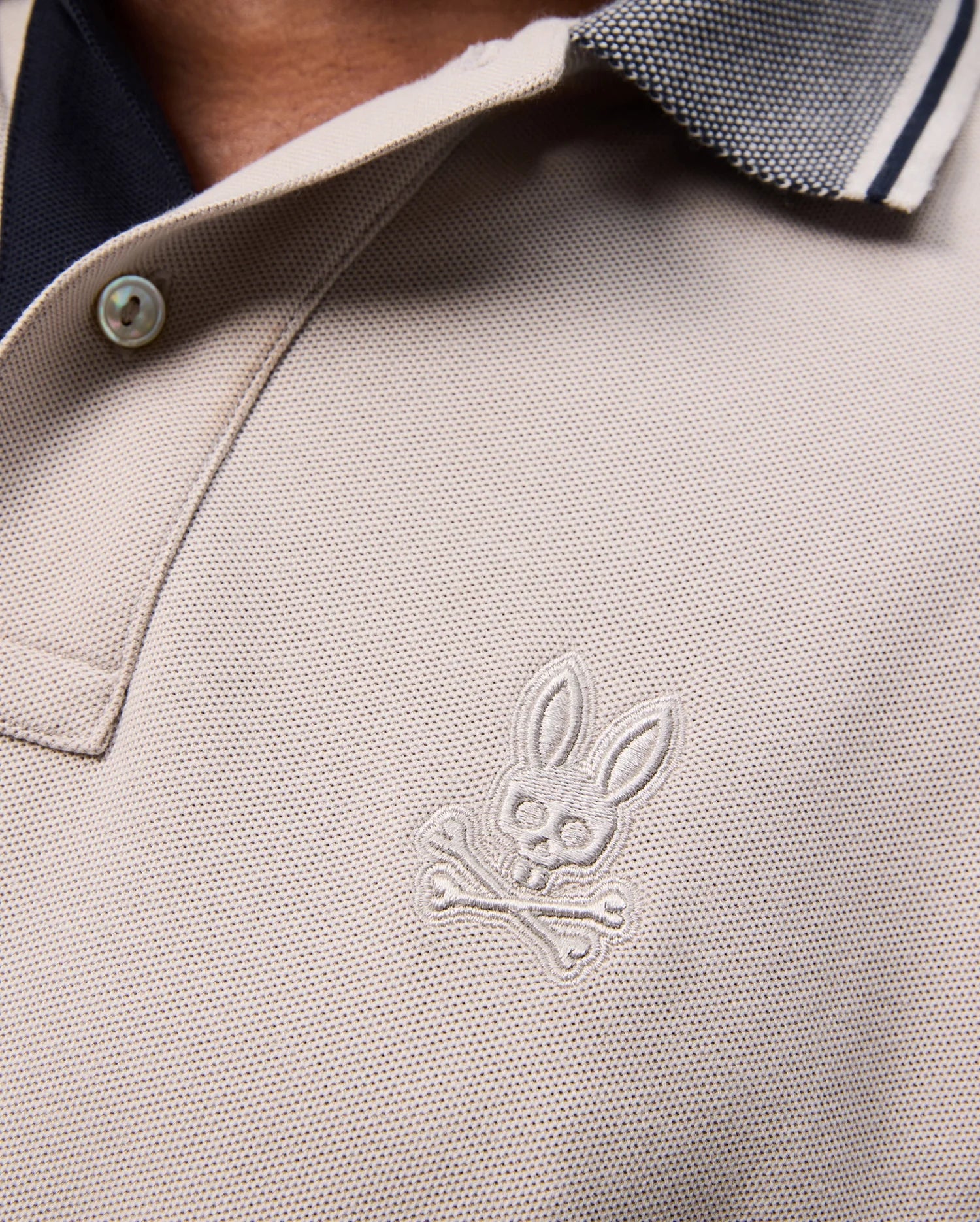 Close-up of the MENS MALTON PIQUE POLO SHIRT - B6K950D200 by Psycho Bunny, crafted from beige Pima cotton with a buttoned collar and black trim. This polo features a white embroidered Psycho Bunny skull and crossbones logo with rabbit ears on the chest, showcasing a subtle diamond pique pattern for added texture.