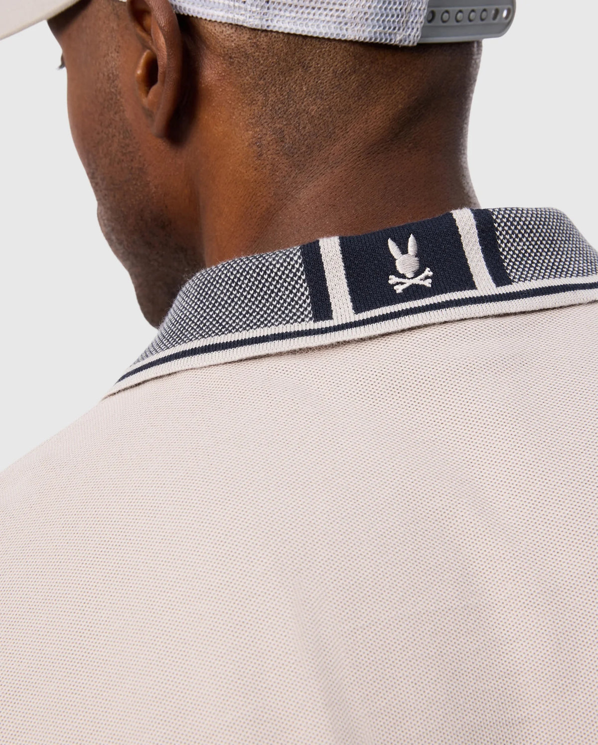Close-up of a man wearing a Psycho Bunny MENS MALTON PIQUE POLO SHIRT - B6K950D200 in beige and a light grey cap. The focus is on the back of the polo shirt's collar, showcasing a white bunny and crossed bones emblem on a navy and white striped background with subtle diamond pique pattern detailing.