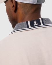 Close-up of a man wearing a Psycho Bunny MENS MALTON PIQUE POLO SHIRT - B6K950D200 in beige and a light grey cap. The focus is on the back of the polo shirt's collar, showcasing a white bunny and crossed bones emblem on a navy and white striped background with subtle diamond pique pattern detailing.