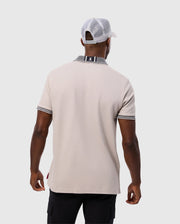 A man seen from the back is wearing a Psycho Bunny MENS MALTON PIQUE POLO SHIRT (B6K950D200) made of beige Pima cotton, featuring a black and white striped collar and sleeve cuffs. He pairs it with black pants, and has a white mesh baseball cap on backward. The background is plain and light gray.