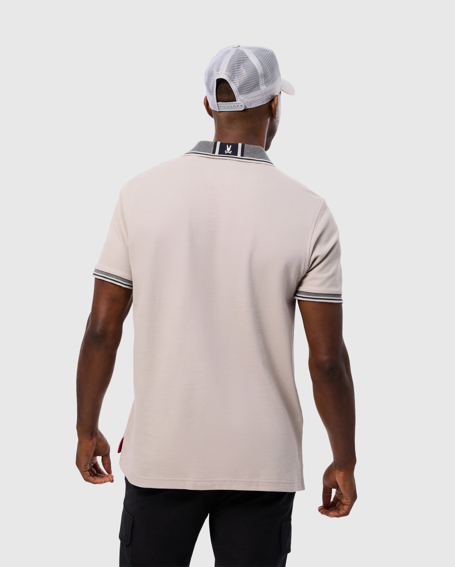 A man seen from the back is wearing a Psycho Bunny MENS MALTON PIQUE POLO SHIRT (B6K950D200) made of beige Pima cotton, featuring a black and white striped collar and sleeve cuffs. He pairs it with black pants, and has a white mesh baseball cap on backward. The background is plain and light gray.