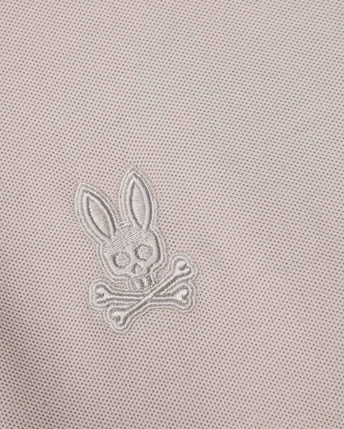 Close-up of the Pima cotton fabric on the Psycho Bunny MENS MALTON PIQUE POLO SHIRT - B6K950D200, showcasing an embroidered design featuring a skull with bunny ears above two crossed bones. The embroidery blends seamlessly into the diamond pique pattern, creating a subtle yet distinctive Psycho Bunny signature.