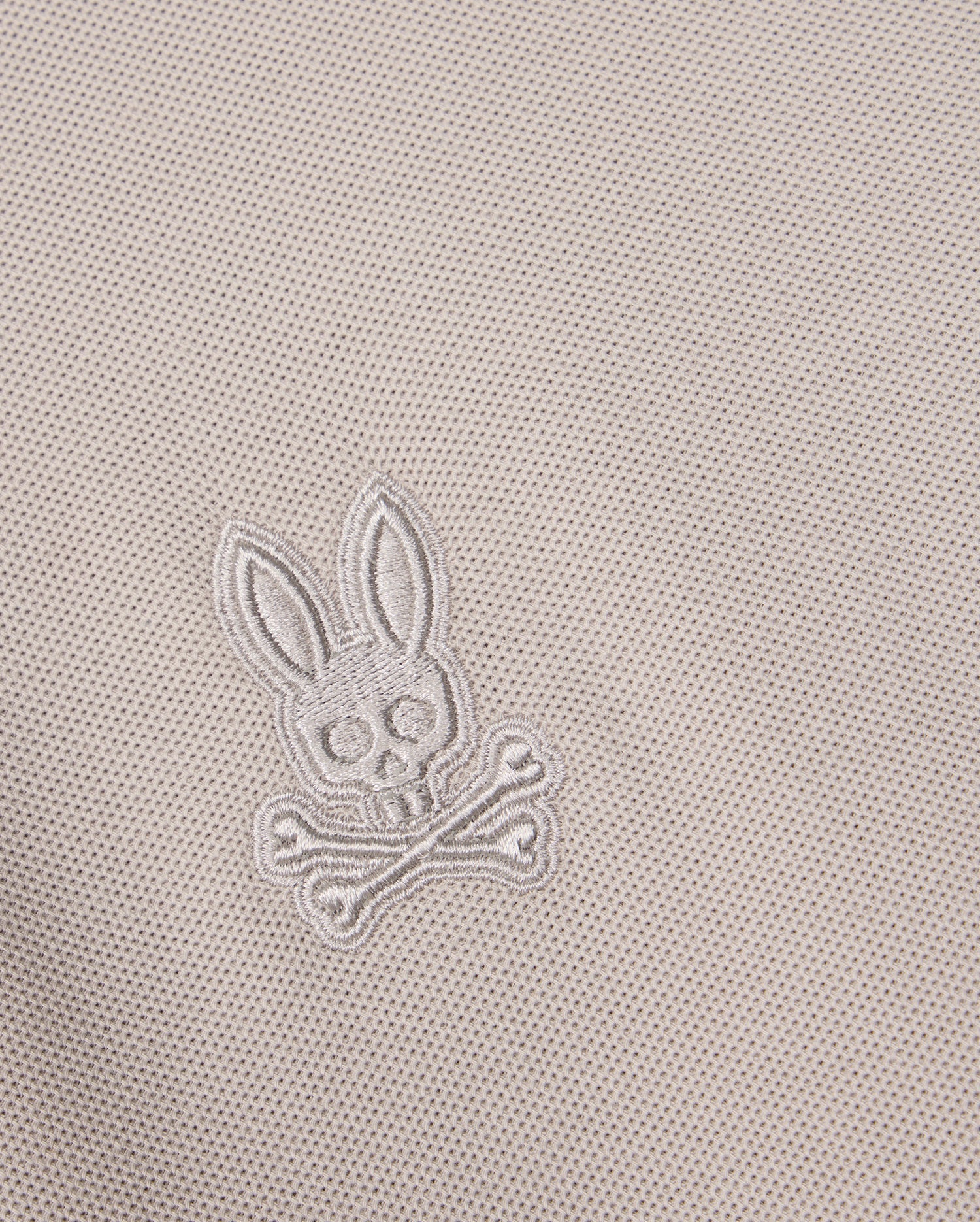 Close-up of the Pima cotton fabric on the Psycho Bunny MENS MALTON PIQUE POLO SHIRT - B6K950D200, showcasing an embroidered design featuring a skull with bunny ears above two crossed bones. The embroidery blends seamlessly into the diamond pique pattern, creating a subtle yet distinctive Psycho Bunny signature.