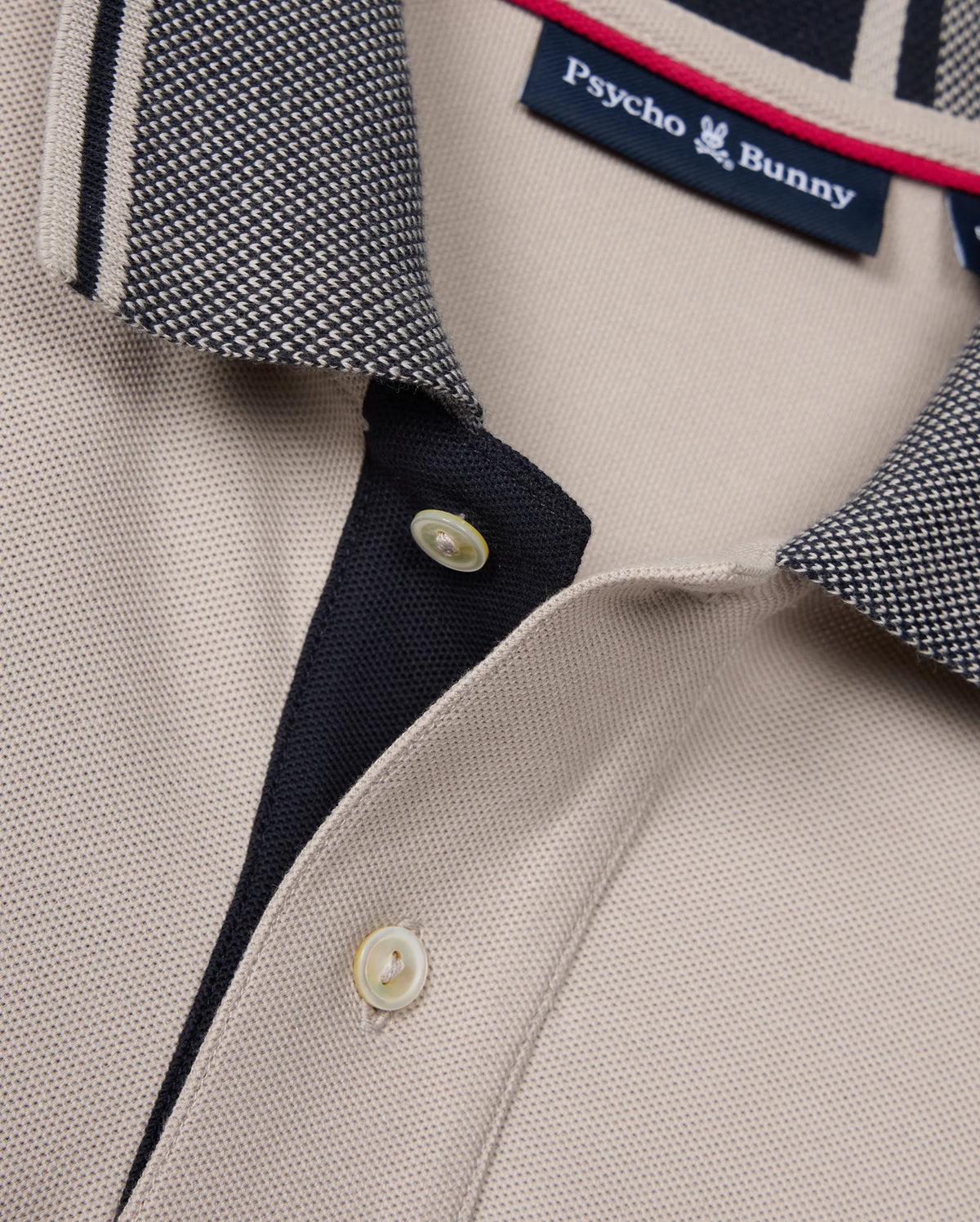 Close-up of the beige Psycho Bunny Mens Malton Pique Polo Shirt (B6K950D200) showcasing a black and white textured collar. The polo features a black placket with two white buttons and a visible Psycho Bunny tag inside the collar. The fabric has a subtle diamond pique pattern, giving it a textured knit appearance.