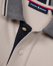 Close-up of the beige Psycho Bunny Mens Malton Pique Polo Shirt (B6K950D200) showcasing a black and white textured collar. The polo features a black placket with two white buttons and a visible Psycho Bunny tag inside the collar. The fabric has a subtle diamond pique pattern, giving it a textured knit appearance.