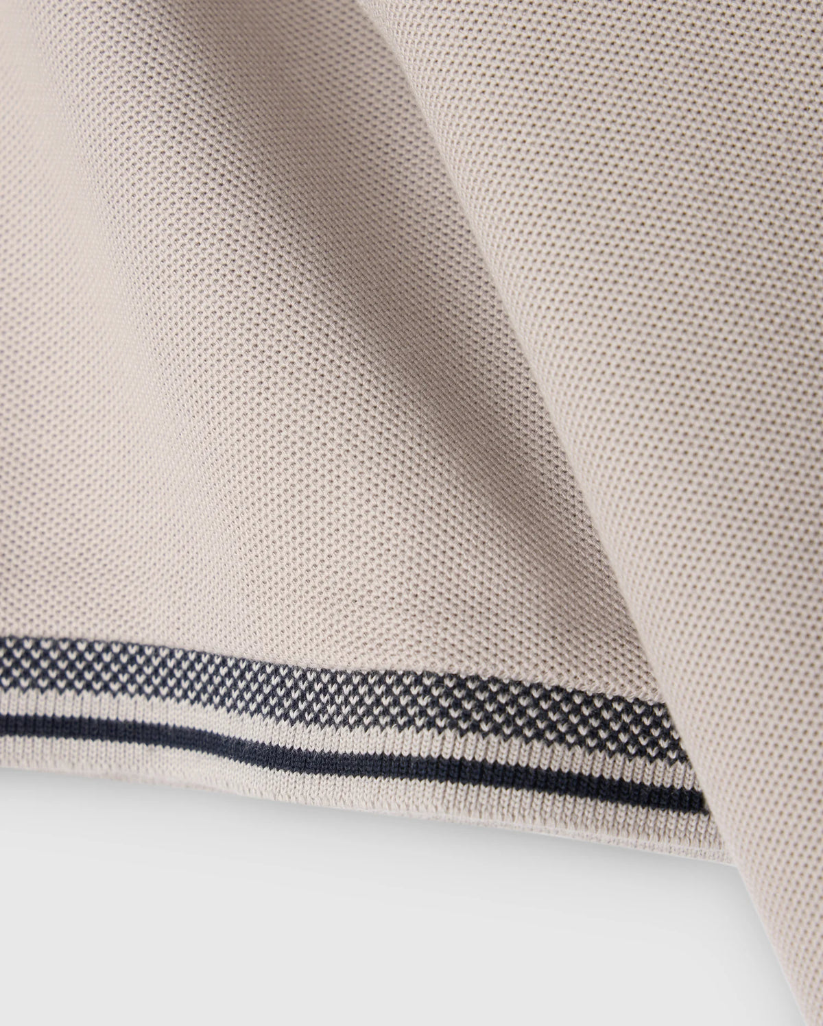 Close-up of a piece of beige fabric with a smooth, textured surface. The fabric edge, folded over, features a decorative pattern with black and white stitching, reminiscent of the intricate detailing found on the Psycho Bunny MENS MALTON PIQUE POLO SHIRT - B6K950D200.