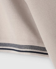 Close-up of a piece of beige fabric with a smooth, textured surface. The fabric edge, folded over, features a decorative pattern with black and white stitching, reminiscent of the intricate detailing found on the Psycho Bunny MENS MALTON PIQUE POLO SHIRT - B6K950D200.