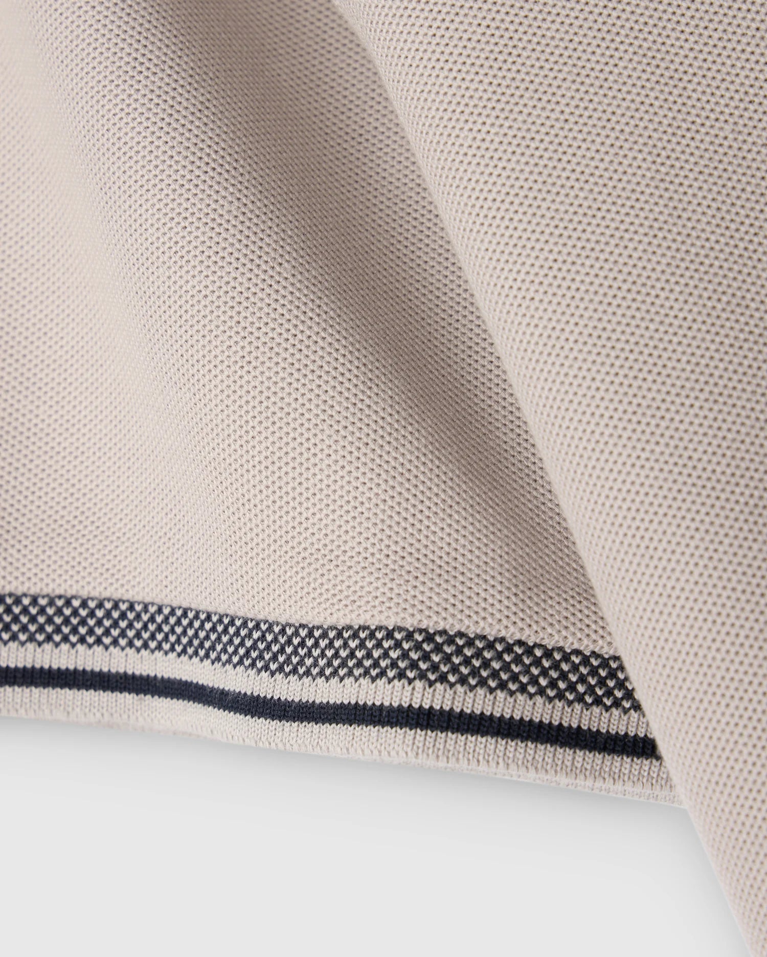 Close-up of a piece of beige fabric with a smooth, textured surface. The fabric edge, folded over, features a decorative pattern with black and white stitching, reminiscent of the intricate detailing found on the Psycho Bunny MENS MALTON PIQUE POLO SHIRT - B6K950D200.