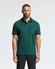 A man with a beard stands against a plain background, wearing the MENS MALTON PIQUE POLO in dark green from Psycho Bunny paired with black pants. He gazes directly at the camera, his arms relaxed by his sides, exuding effortless style and confidence.