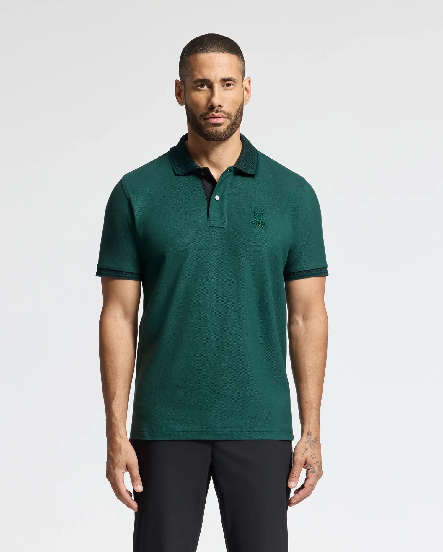 A man with a beard stands against a plain background, wearing the MENS MALTON PIQUE POLO in dark green from Psycho Bunny paired with black pants. He gazes directly at the camera, his arms relaxed by his sides, exuding effortless style and confidence.