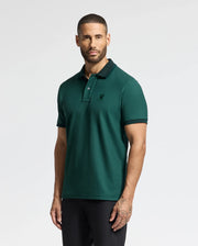 A person with short hair and a beard is wearing the dark green MENS MALTON PIQUE POLO - B6K950D200 by Psycho Bunny, featuring a small emblem on the chest. They are standing against a plain white background, highlighting the lightweight comfort of this stylish garment.