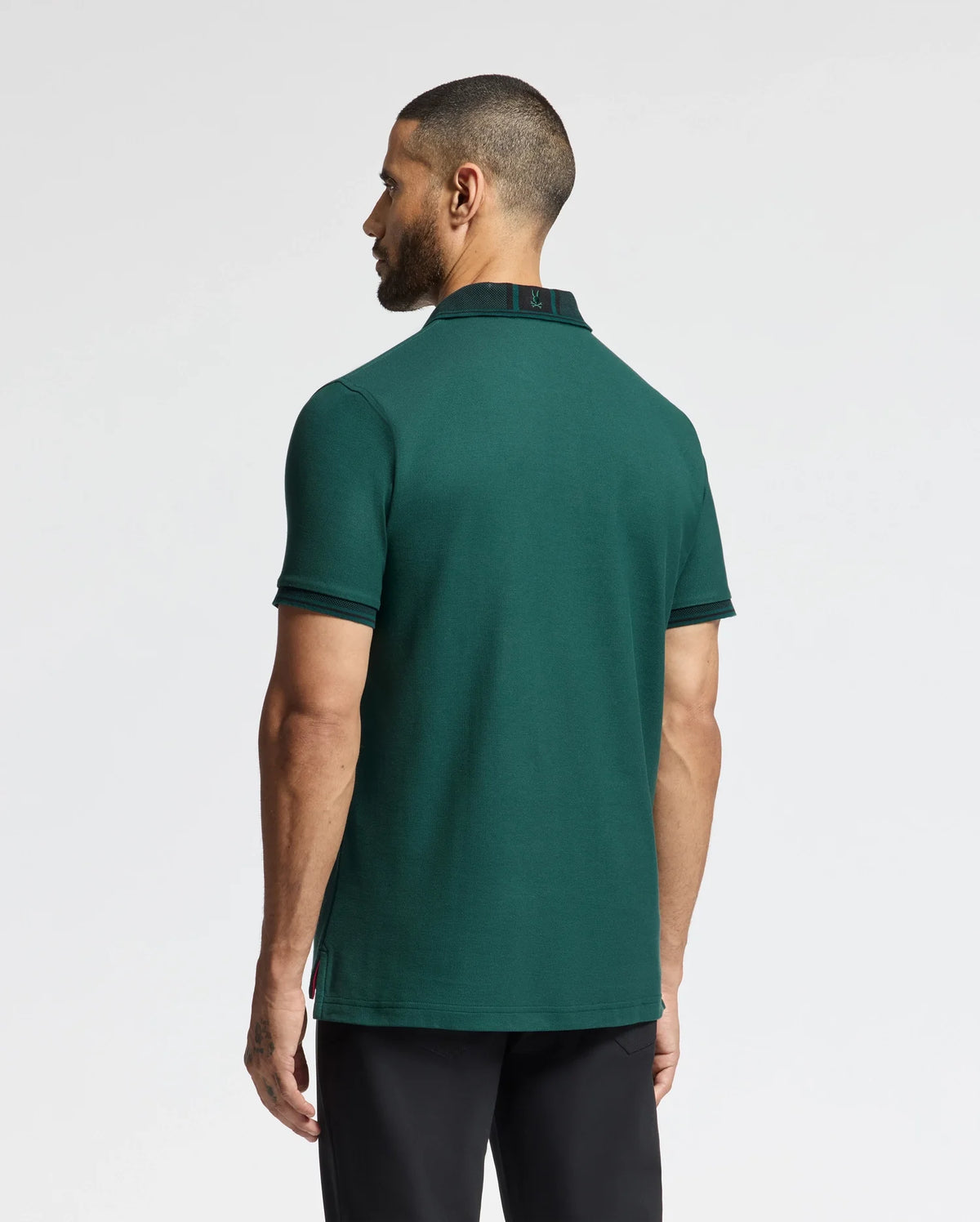 A man with a short haircut stands with his back to the camera, dressed in a dark green Psycho Bunny MENS MALTON PIQUE POLO - B6K950D200 and black pants, set against a plain white background.