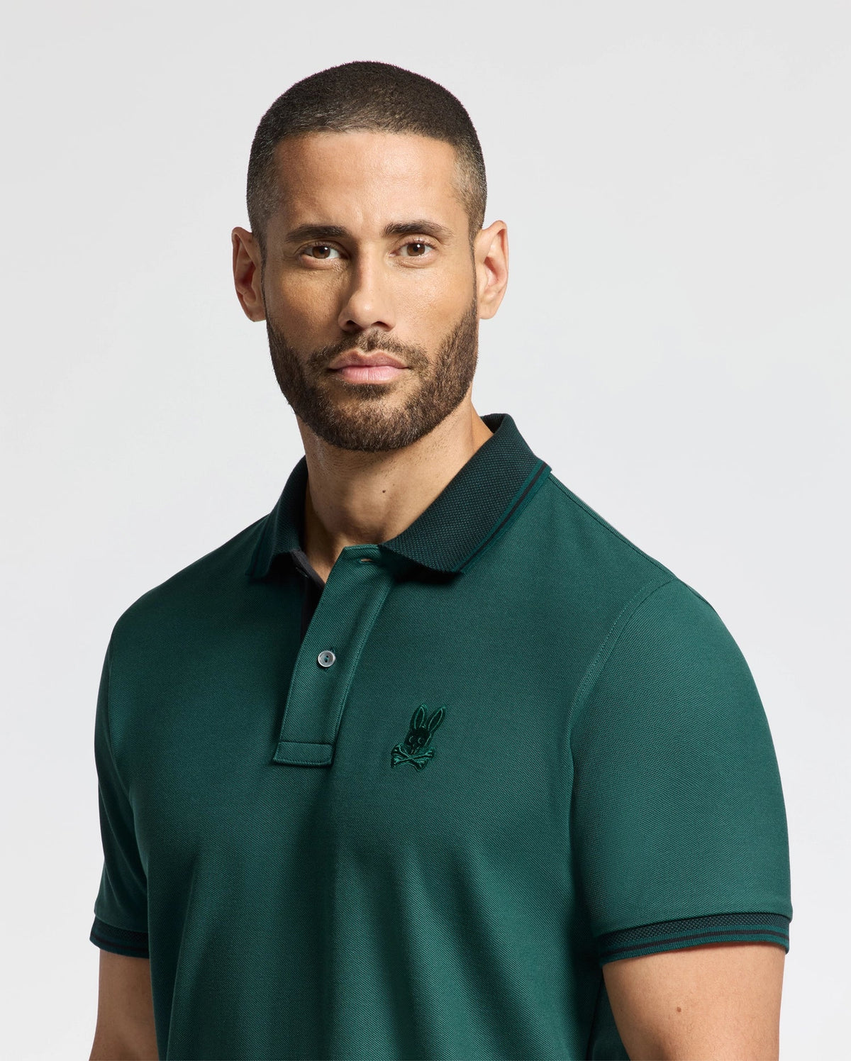 A man with short hair and a beard is wearing Psycho Bunny's MENS MALTON PIQUE POLO - B6K950D200 in dark green, showcasing its high-quality, lightweight design and a subtle embroidered logo on the chest. The plain, light-colored background accentuates the comfort and style of this premium men's attire.