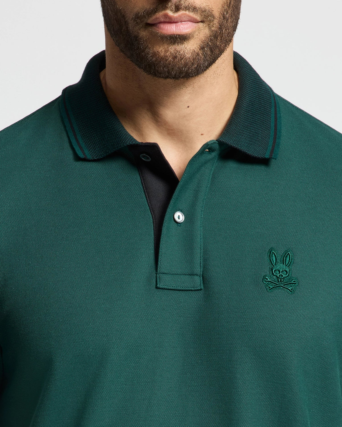 A man wearing the Psycho Bunny MENS MALTON PIQUE POLO - B6K950D200 in dark green with a black inner lining. The lightweight polo boasts a small embroidered emblem of a bunny skull and crossbones on the left side. The image has cropped out part of his head.