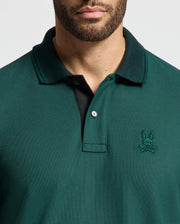 A man wearing the Psycho Bunny MENS MALTON PIQUE POLO - B6K950D200 in dark green with a black inner lining. The lightweight polo boasts a small embroidered emblem of a bunny skull and crossbones on the left side. The image has cropped out part of his head.