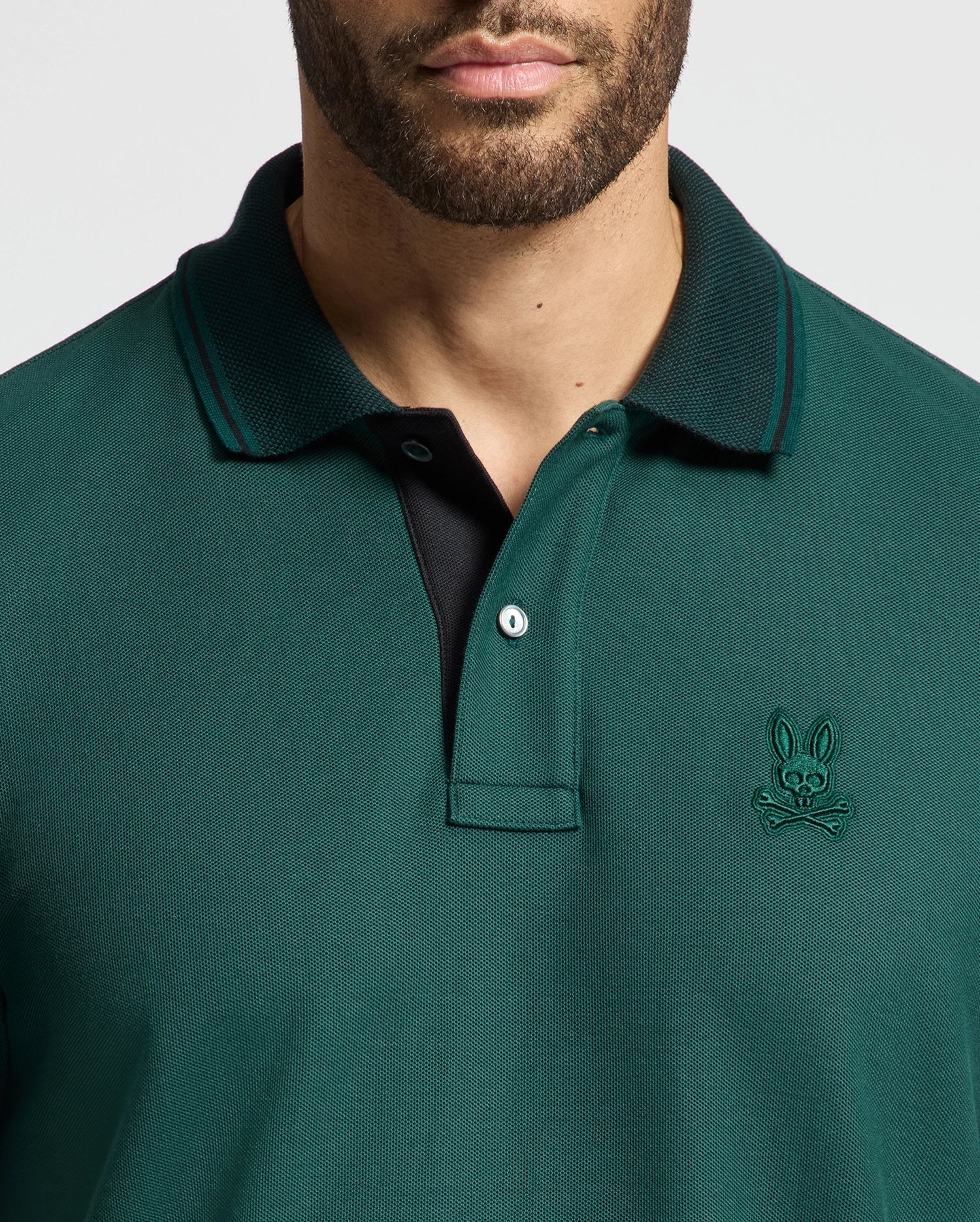 A man wearing the Psycho Bunny MENS MALTON PIQUE POLO - B6K950D200 in dark green with a black inner lining. The lightweight polo boasts a small embroidered emblem of a bunny skull and crossbones on the left side. The image has cropped out part of his head.