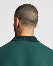 A person with a close-cropped haircut is facing away from the camera, wearing a Mens Malton Pique Polo from Psycho Bunny in dark green. Crafted from lightweight pique fabric, this high-quality men's polo features a small embroidered logo on the back of the collar. The background is light gray.