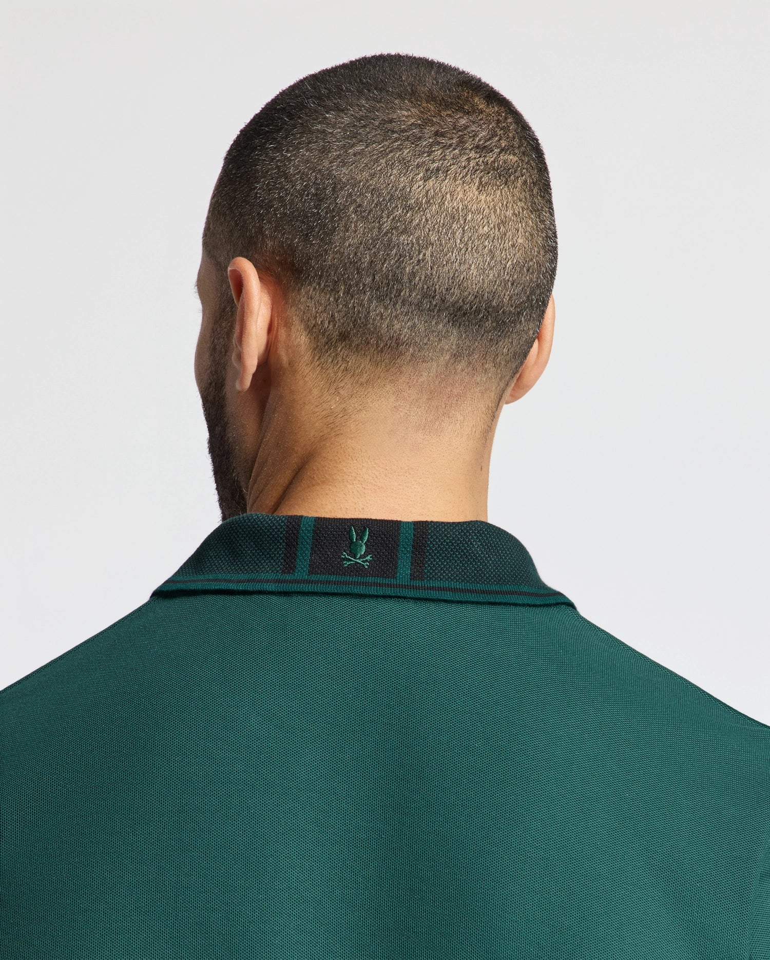 A person with a close-cropped haircut is facing away from the camera, wearing a Mens Malton Pique Polo from Psycho Bunny in dark green. Crafted from lightweight pique fabric, this high-quality men's polo features a small embroidered logo on the back of the collar. The background is light gray.