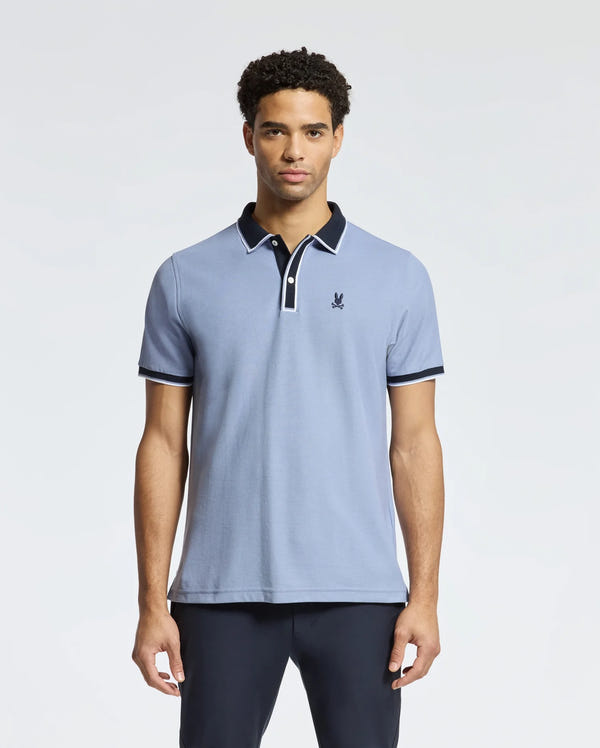 A man wearing the Psycho Bunny MENS KENT PIQUE POLO - B6K951D200 in light blue with a contrasting dark collar and a small embroidered logo on the chest. This pique polo for men, featuring dark trim on the sleeves, is showcased against a plain white background.