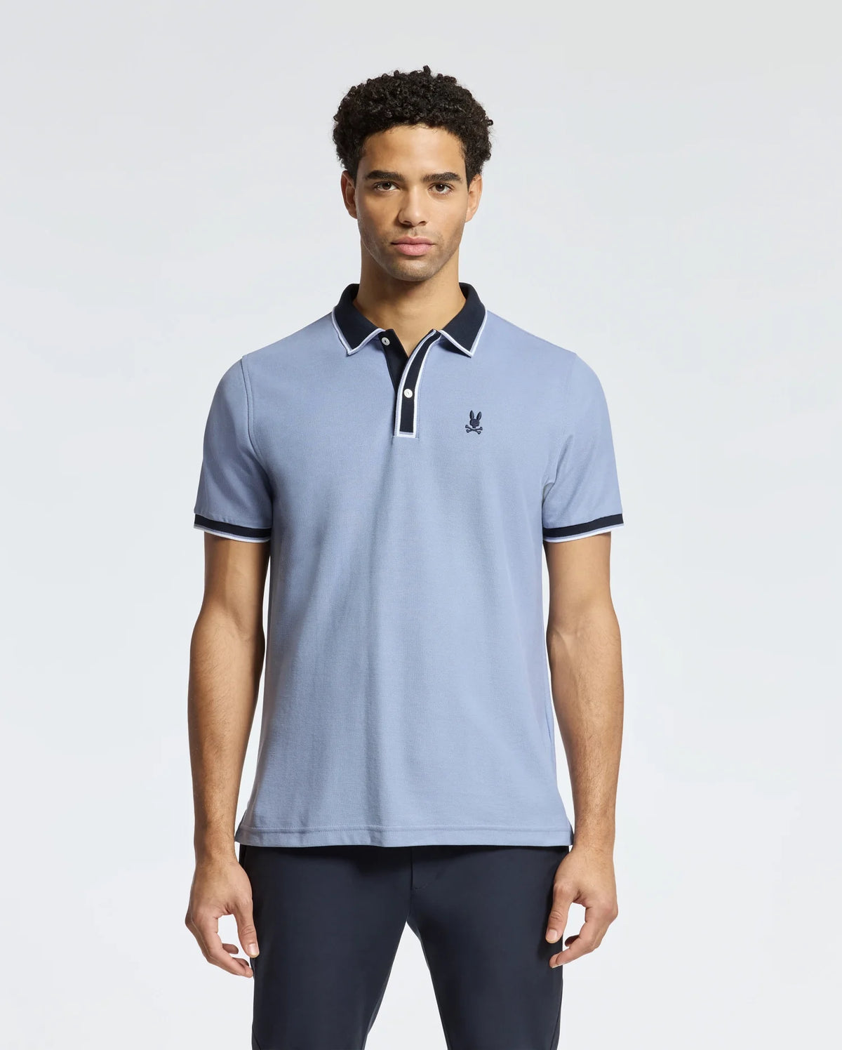 A man wearing the Psycho Bunny MENS KENT PIQUE POLO - B6K951D200 in light blue with a contrasting dark collar and a small embroidered logo on the chest. This pique polo for men, featuring dark trim on the sleeves, is showcased against a plain white background.