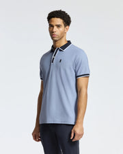 A man sporting the Psycho Bunny MENS KENT PIQUE POLO in light blue with dark trim on the collar and sleeves stands against a plain background. This polo showcases a discreet embroidered logo on the chest. He has short curly hair and is glancing slightly to the side.