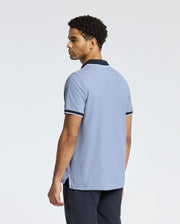 A man stands with his back to the camera wearing a Psycho Bunny MENS KENT PIQUE POLO - B6K951D200, featuring a light blue color accented by dark trim on the collar and sleeves. His short, curly hair harmonizes perfectly with his classic dark pants. The simple white background enhances the timeless elegance of his outfit.