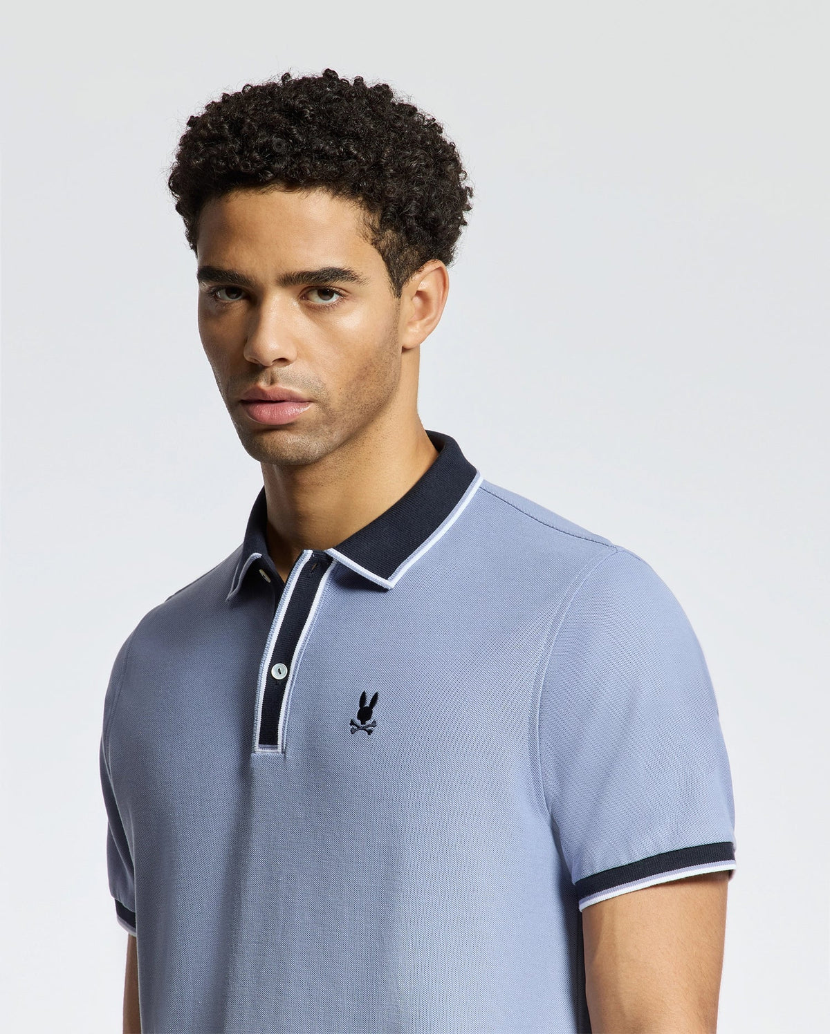 A man wearing the Psycho Bunny's MENS KENT PIQUE POLO (B6K951D200), featuring a dark collar and a small embroidered logo, stands against a plain background. The light blue color of the premium polo made from Pima cotton complements his short, curly hair and neutral expression.