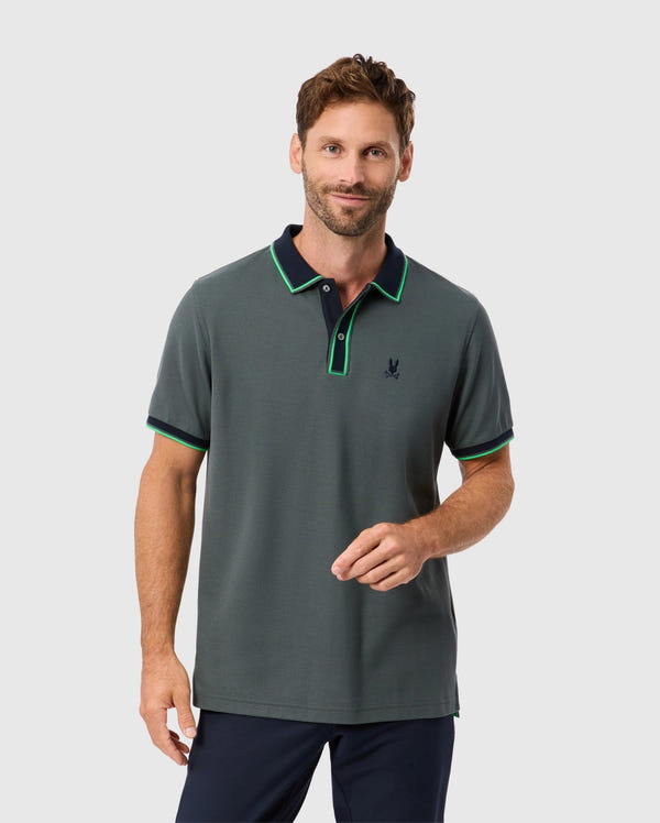 A man with short hair and a beard is wearing a stylish MENS KENT PIQUE POLO SHIRT - B6K951D200 by Psycho Bunny. The shirt features a chic dark green color with a black and green collar, made from premium Pima cotton. He pairs it with dark trousers and stands against a light gray background, looking forward with a slight smile while holding his fingers together.