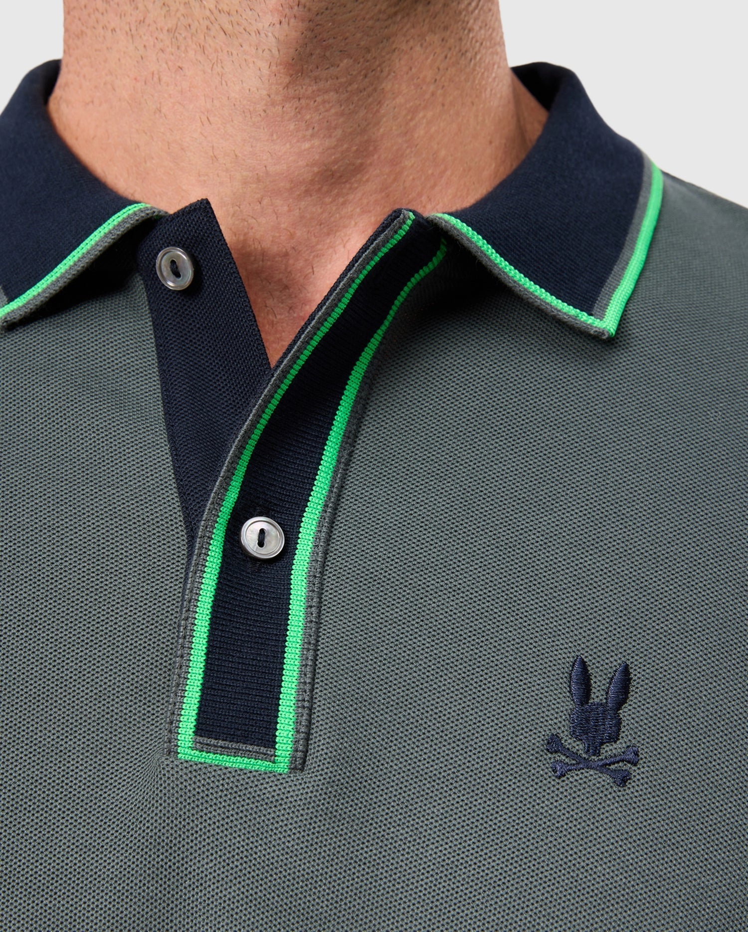 Close-up of a person wearing the MENS KENT PIQUE POLO SHIRT - B6K951D200 from Psycho Bunny, showcasing its stylish Pima cotton fabric in a gray hue. The polo features a dark navy collar and green trim, with an embroidered logo of a stylized skull and rabbit ears on the chest and two buttons at the collar.