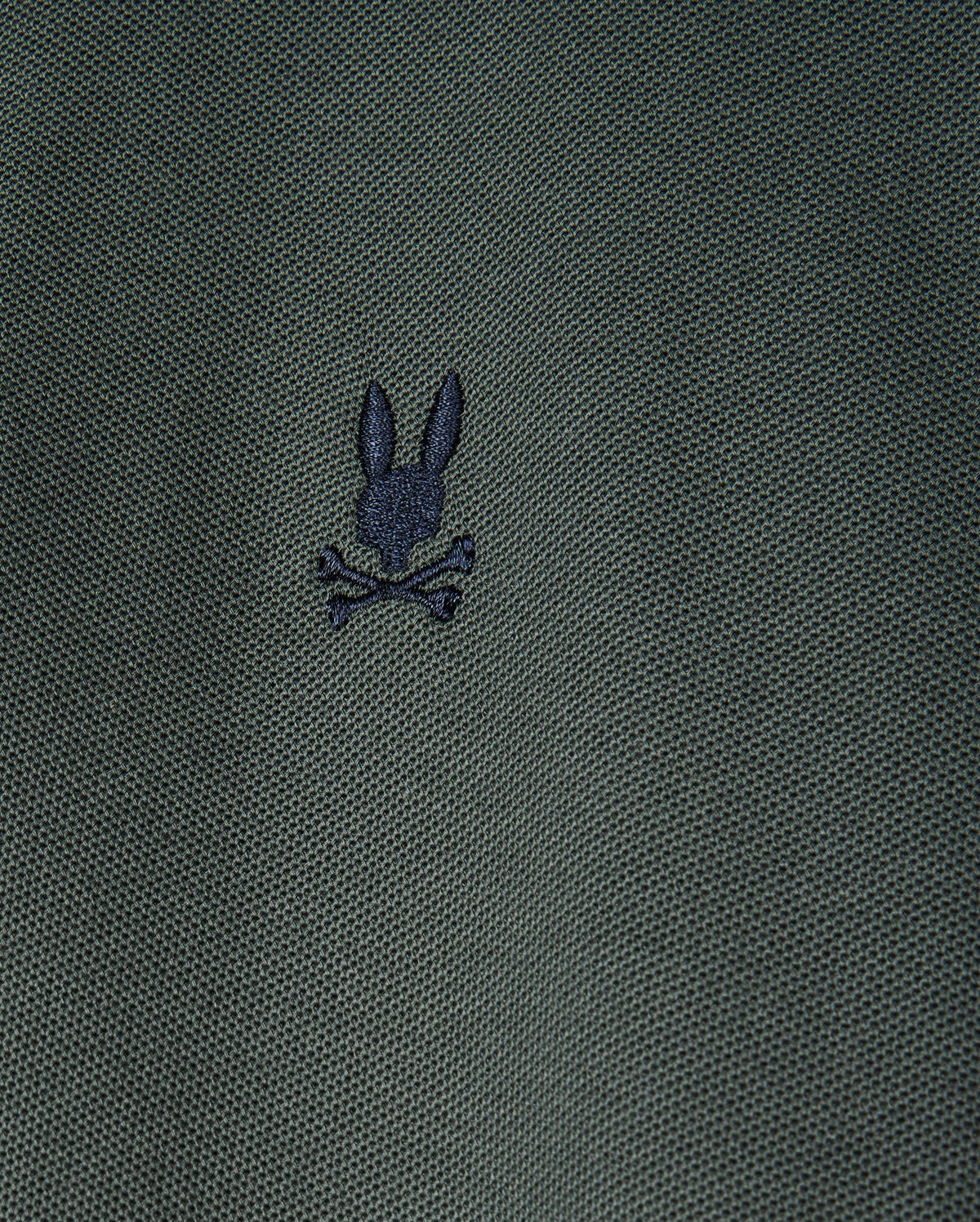 A close-up image of the textured dark green fabric from the Men's Kent Pique Polo Shirt by Psycho Bunny showcases a small embroidered logo, featuring a creative twist on the classic skull and crossbones design with a black silhouette of a rabbit's head above crossed bones.