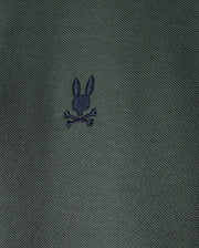 A close-up image of the textured dark green fabric from the Men's Kent Pique Polo Shirt by Psycho Bunny showcases a small embroidered logo, featuring a creative twist on the classic skull and crossbones design with a black silhouette of a rabbit's head above crossed bones.