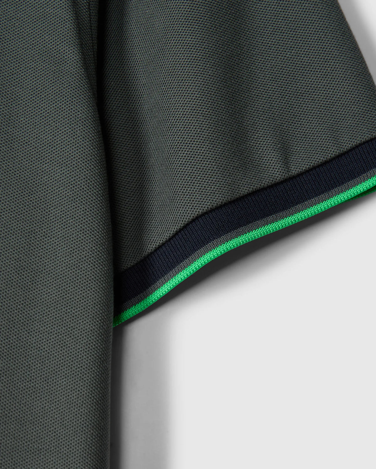 Close-up of the sleeve on a stylish MENS KENT PIQUE POLO SHIRT - B6K951D200 by Psycho Bunny. The dark green Pima cotton fabric is textured for added depth. The hem showcases a thin green stripe alongside a slightly thicker black stripe, providing contrast against the solid light gray background.