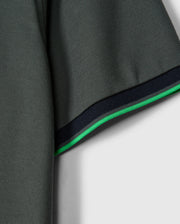 Close-up of the sleeve on a stylish MENS KENT PIQUE POLO SHIRT - B6K951D200 by Psycho Bunny. The dark green Pima cotton fabric is textured for added depth. The hem showcases a thin green stripe alongside a slightly thicker black stripe, providing contrast against the solid light gray background.