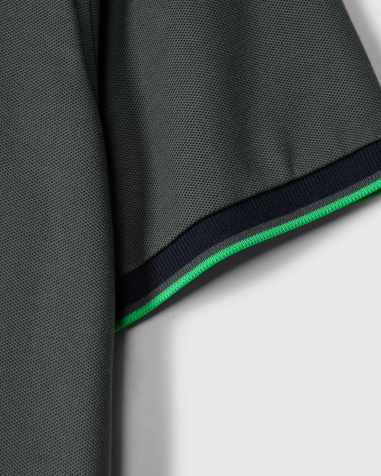 Close-up of the sleeve on a stylish MENS KENT PIQUE POLO SHIRT - B6K951D200 by Psycho Bunny. The dark green Pima cotton fabric is textured for added depth. The hem showcases a thin green stripe alongside a slightly thicker black stripe, providing contrast against the solid light gray background.