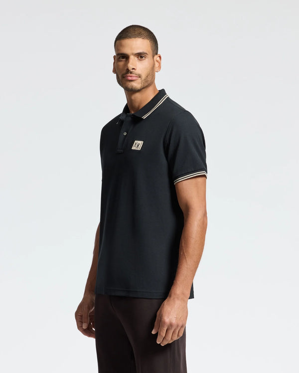 A man with short hair is wearing the Psycho Bunny MENS MONROE PIQUE POLO - B6K964D200, a black Pima cotton polo featuring a small logo on the chest and Mother-of-Pearl buttons. He stands against a plain white background, facing slightly to the left with his hands at his sides.