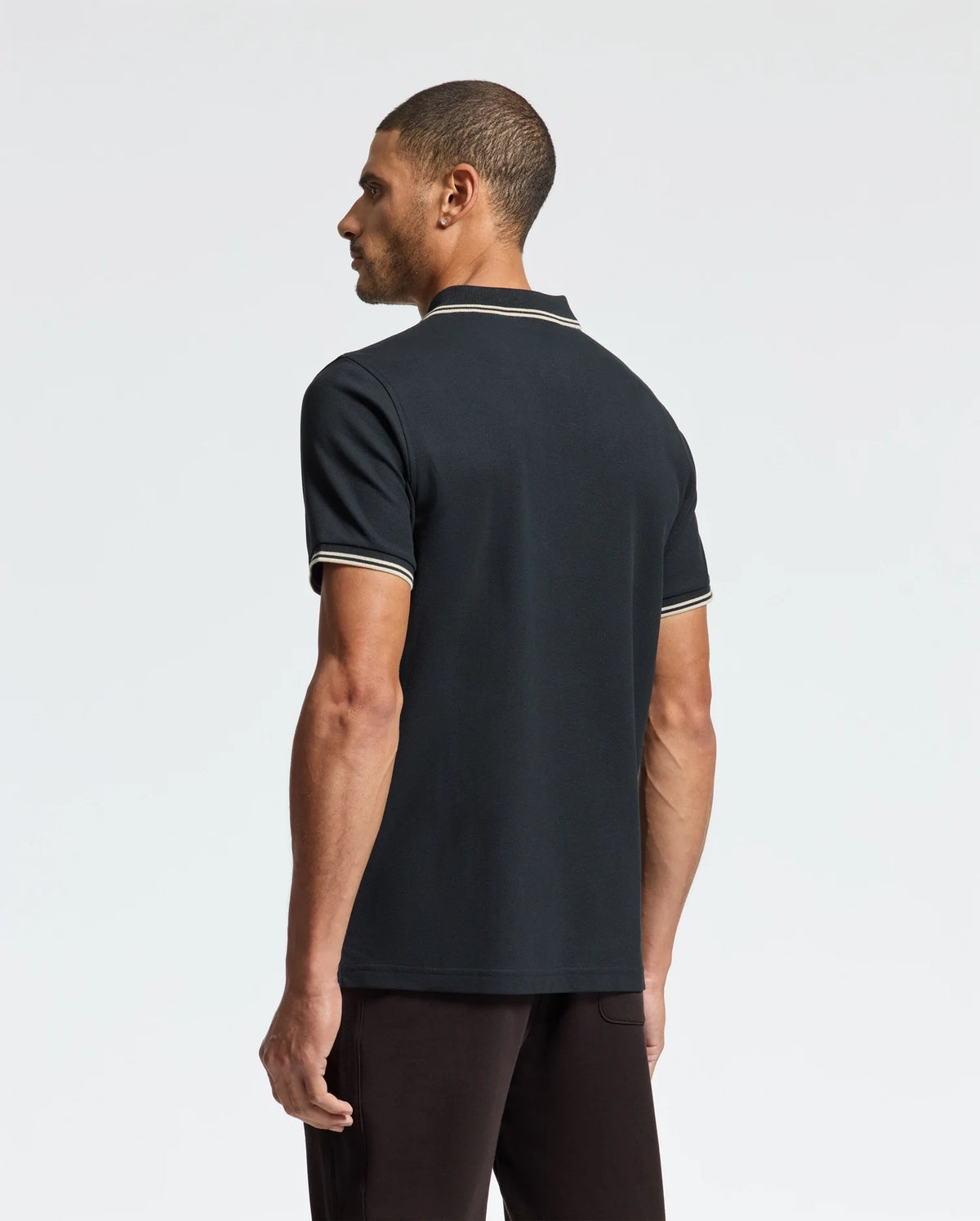 A man wearing the Psycho Bunny MENS MONROE PIQUE POLO - B6K964D200, a regular fit polo crafted from Pima cotton, stands facing away while looking to the side. The polo is stylishly detailed with white trim on the collar and sleeves and adorned with elegant Mother-of-Pearl buttons. He pairs it with dark pants against a plain light background.