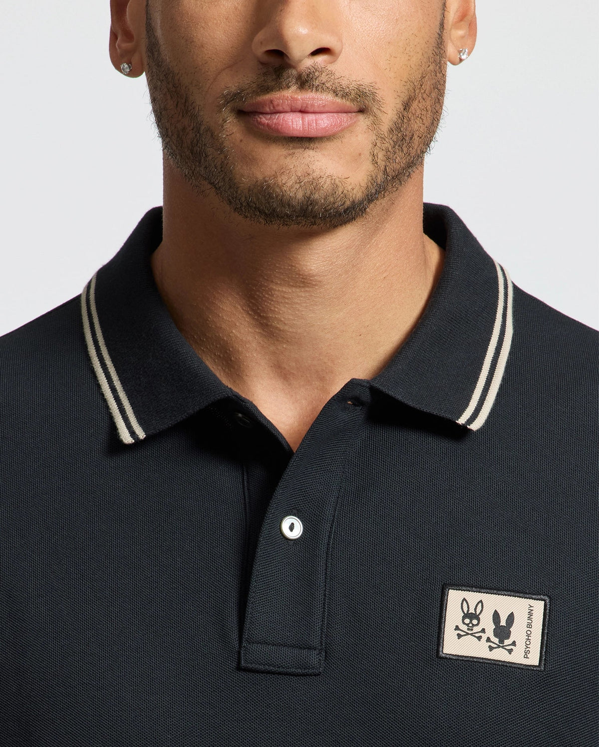 A close-up of a man wearing a Psycho Bunny MENS MONROE PIQUE POLO in black, embellished with white trim and Mother-of-Pearl buttons. The polo shirt displays a patch featuring two rabbits on the chest. He has facial hair, wears small earrings, and is positioned against a simple, light-colored backdrop.