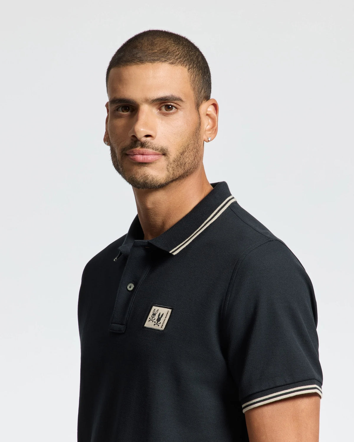 A man with short hair and a beard is wearing the Psycho Bunny MENS MONROE PIQUE POLO - B6K964D200, a dark regular fit polo shirt crafted from Pima cotton and adorned with a chest logo. The Mother-of-Pearl buttons add an elegant touch as he gazes slightly to the side against a plain background.