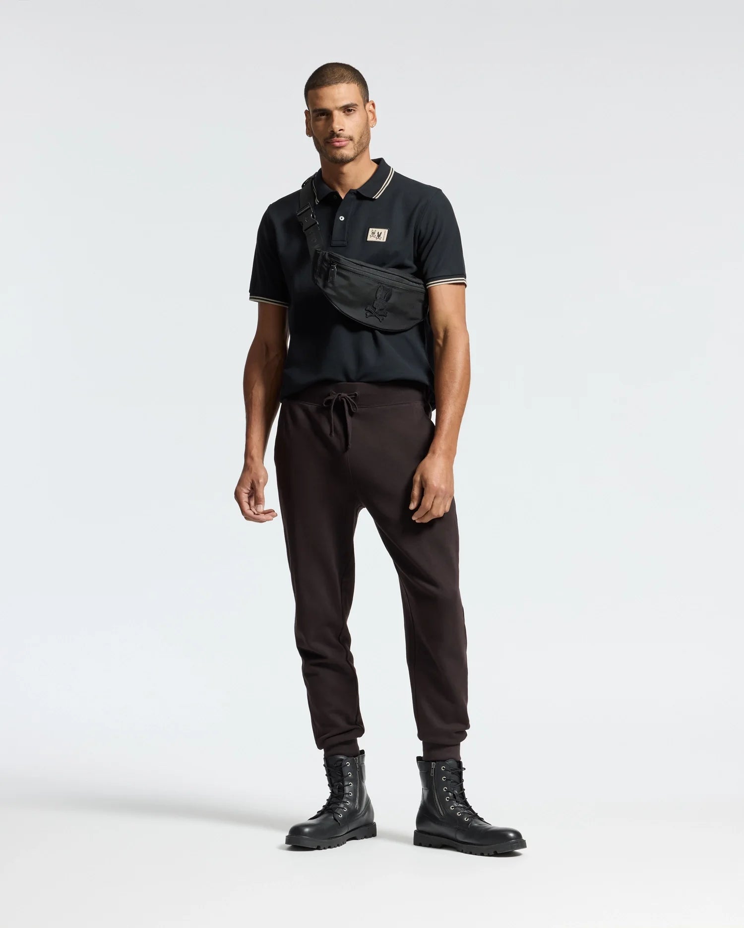 A man stands against a simple backdrop wearing a black Monroe Pique Polo from Psycho Bunny, paired with dark brown jogger pants and black lace-up boots. Draped across his chest is a black crossbody bag featuring Mother-of-Pearl buttons. His expression is neutral, while his arms rest easily by his sides.