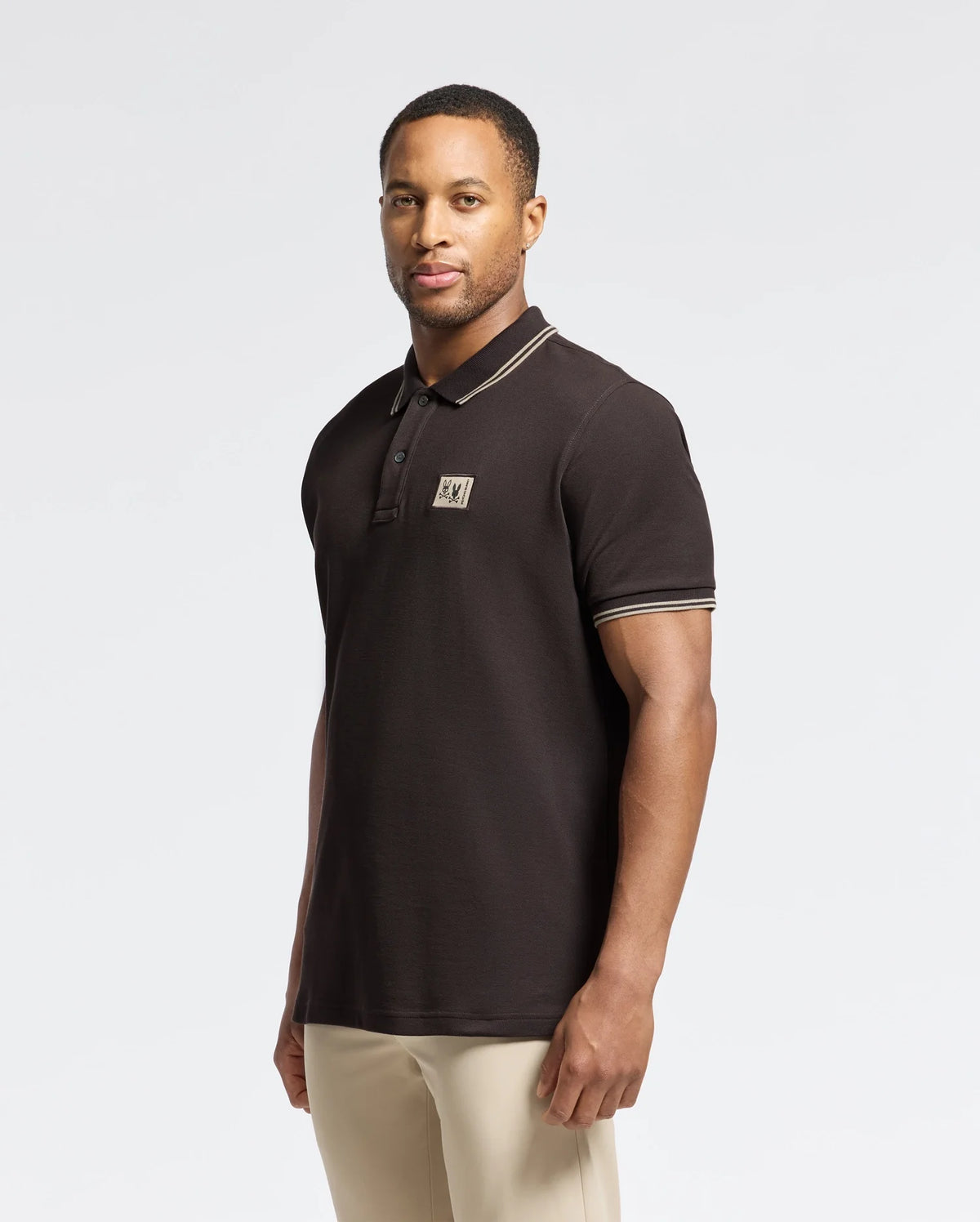 A man with short hair wears the Psycho Bunny MENS MONROE PIQUE POLO - B6K964D200, a black Pima cotton polo featuring contrasting trim and Mother-of-Pearl buttons, against a plain background. A small logo is displayed on the chest of this Big and Tall-friendly garment. He pairs it with light-colored pants.
