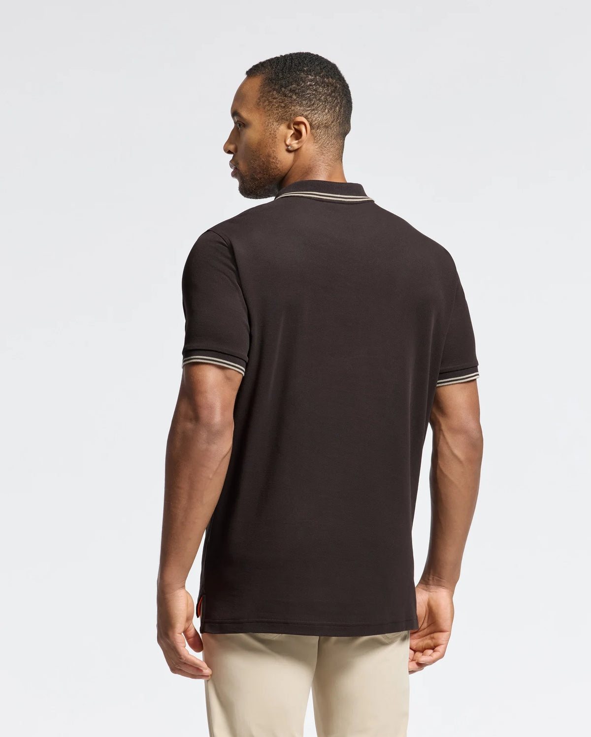 A man is standing with his back to the camera, wearing a Psycho Bunny MENS MONROE PIQUE POLO - B6K964D200 in black, which features mother-of-pearl buttons, paired with beige pants. The background is a neutral light gray.