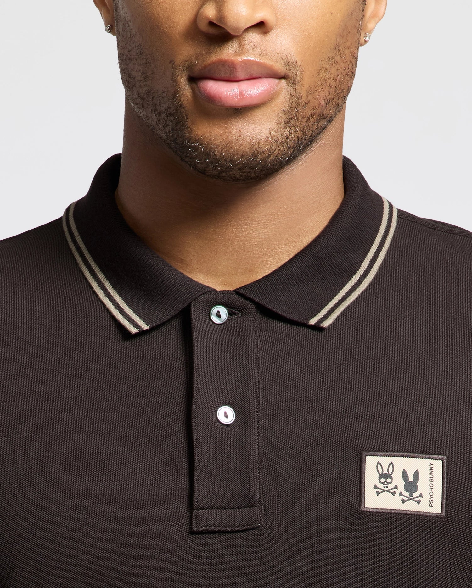 A person is wearing the Psycho Bunny MENS MONROE PIQUE POLO - B6K964D200, a dark Pima cotton polo in Big and Tall sizes, featuring double stripes on the collar and a patch with two cartoon skulls on the left side. The background color of the shirt is light gray.