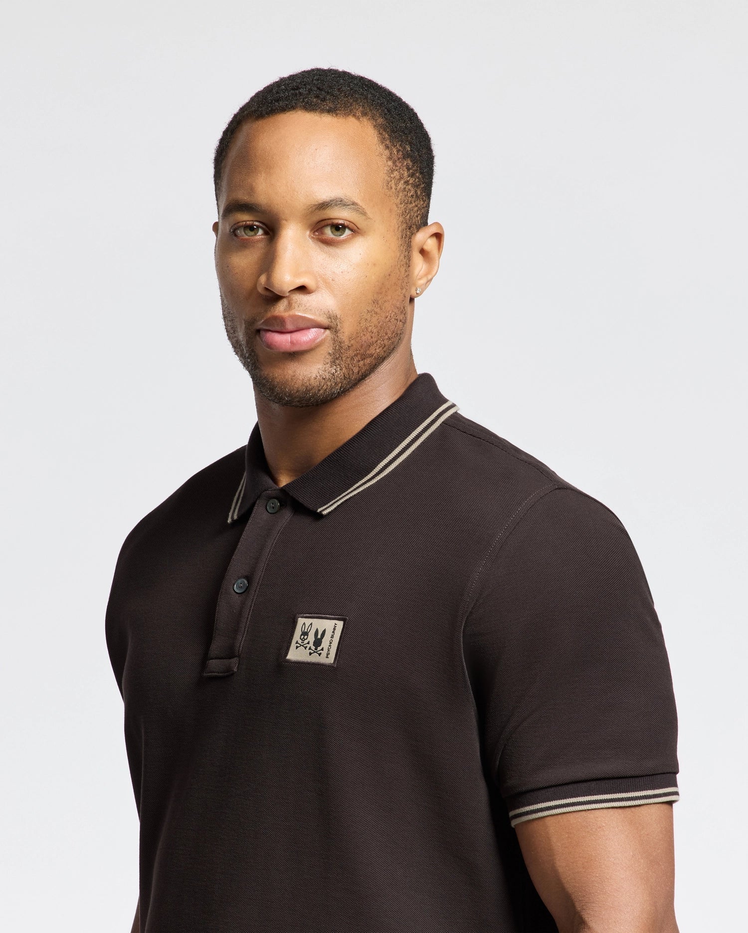 A person wearing the MENS MONROE PIQUE POLO from Psycho Bunny, in black Pima cotton featuring Mother-of-Pearl buttons and a subtle emblem on the chest, stands against a plain background, looking forward.