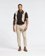 A man wearing the Psycho Bunny Monroe Pique Polo - B6K964D200, made from Pima cotton with mother-of-pearl buttons, stands against a plain background in beige pants. He has a sweater draped around his shoulders and wears brown shoes. His left arm is relaxed by his side, while his right hand is partially in his pocket.