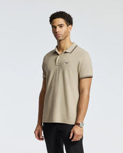A man is standing against a plain white background dressed in the MENS MONROE PIQUE POLO from Psycho Bunny, featuring a regular fit and crafted from beige Pima cotton, proudly made in Peru. The polo displays a logo on the chest and is paired with black pants. He has short curly hair and wears a watch on his left wrist.