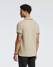 A man with curly hair, wearing the Psycho Bunny Men's Monroe Pique Polo in beige with dark trim and regular fit black pants, stands facing away while glancing to the side. The plain light gray background highlights the quality craftsmanship of this polo shirt, made in Peru.