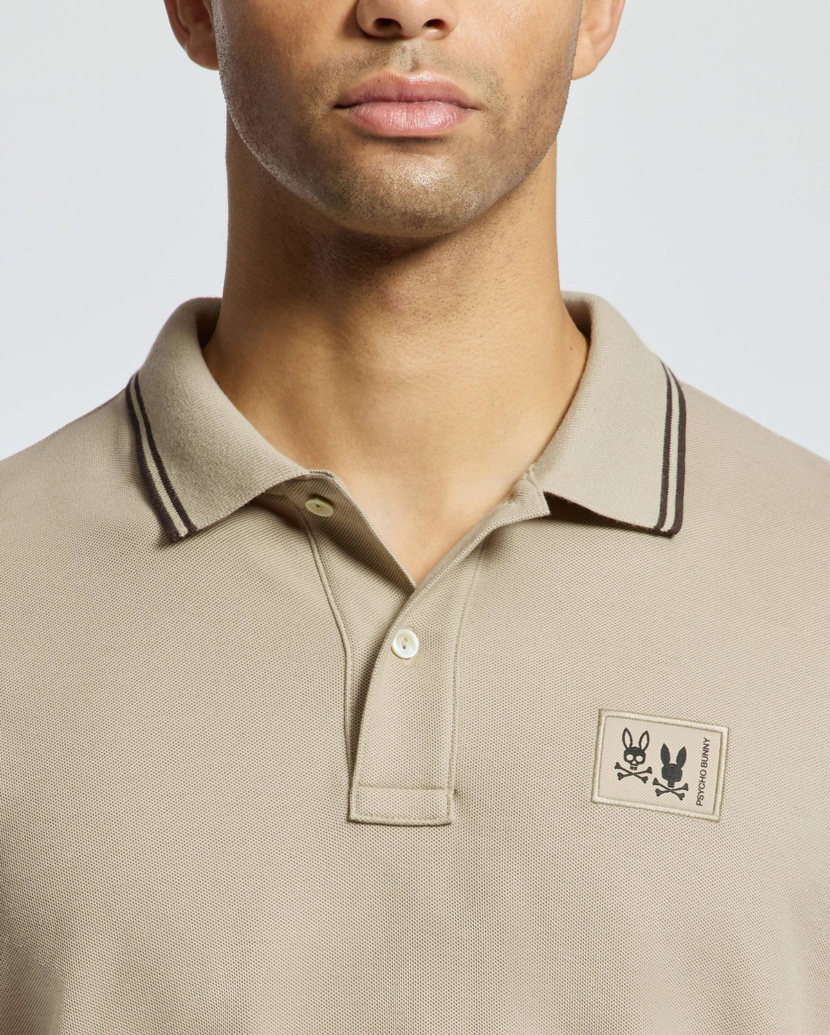 A person wearing a beige Monroe Pique Polo by Psycho Bunny, featuring dark trim on the collar. The shirt, crafted from Pima cotton in Peru, showcases a small floral patch and logo on the left side. The background is a neutral light gray.